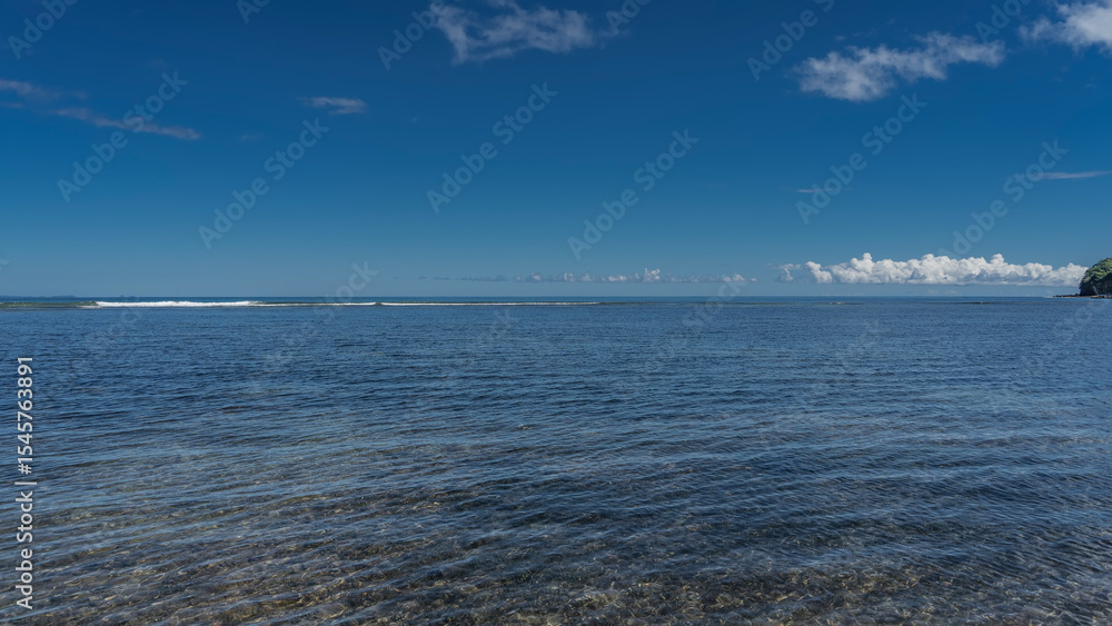 Obraz premium Beautiful calm seascape. Ripples on the surface of the blue ocean. The bottom is visible through the clear water. Foam waves in the distance. Azure sky, white clouds. Copy space. Madagascar. Nosy Be 