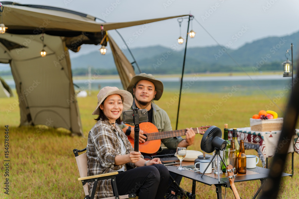 Asian couple camping in a vast grassland during sunset. The woman smiles and takes a selfie while the man plays the guitar. They spend a relaxing time in the peaceful nature. Select a focus point Asian couple camping in a vast grassland during sunset. The woman smiles and takes a selfie while the man plays the guitar. They spend a relaxing time in the peaceful nature. Select a focus point