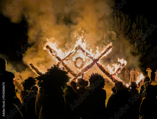 People in traditional Latvian costumes and wreaths watching burning structure shaped as latvian traditional austras tree ornament at Midsummer celebration