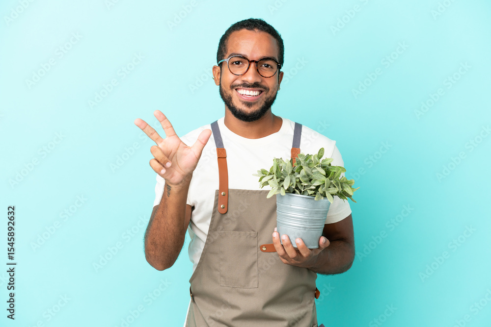Gardener latin man holding a plant isolated on blue background happy and counting three with fingers Gardener latin man holding a plant isolated on blue background happy and counting three with fingers