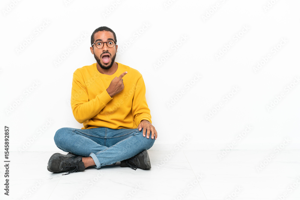 Young Ecuadorian man sitting on the floor isolated on white wall surprised and pointing side Young Ecuadorian man sitting on the floor isolated on white wall surprised and pointing side