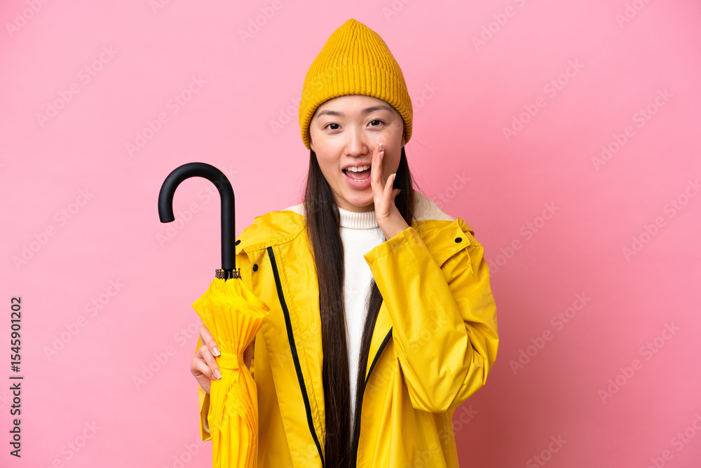 Young Chinese woman with rainproof coat and umbrella isolated on pink background shouting with mouth wide open Young Chinese woman with rainproof coat and umbrella isolated on pink background shouting with mouth wide open