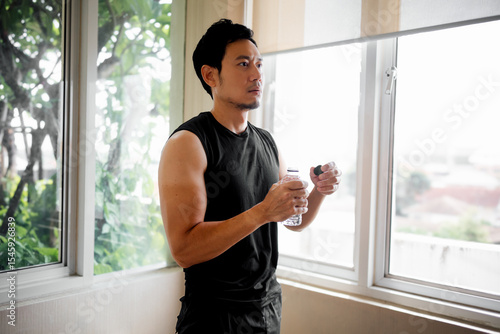 Billede på lærred Asian man holding water bottle and looking out window