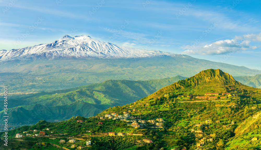panoramic view of a national park with green mountains with palace and great white snowy top of a mountain on a blue cloudy sky background. Panorama of Taormina, Sicily, Italy panoramic view of a national park with green mountains with palace and great white snowy top of a mountain on a blue cloudy sky background. Panorama of Taormina, Sicily, Italy
