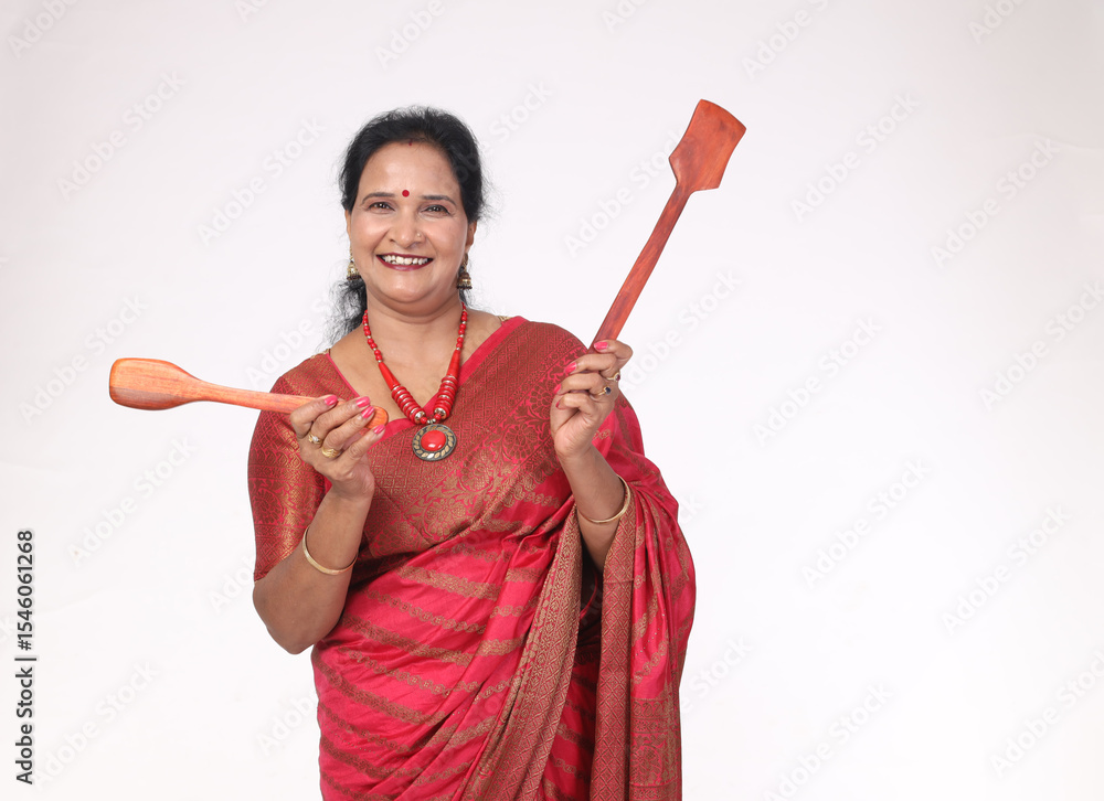 Fototapeta premium Indian happy woman holding wooden spatula. She wearing traditional saree showing kitchen items and home appliances products for cooking food. while standing isolated over white background