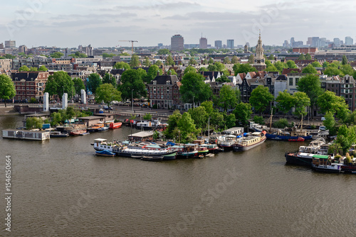 An expansive aerial view of an Amsterdam canal or river, dotted with boats and flanked by tree-lined banks and distant city buildings