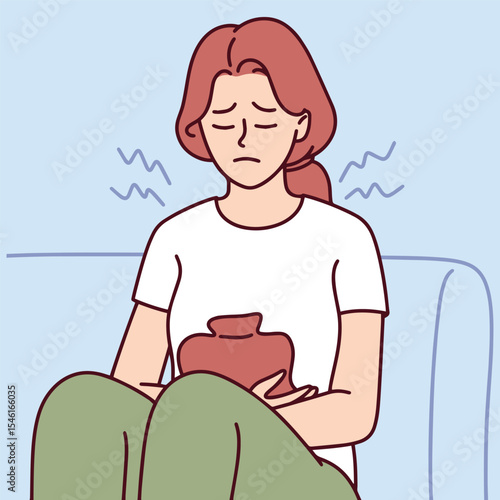 Young woman holds heating pad against stomach while sitting curled up on sofa. She suffers from menstrual cramps and painful period symptoms