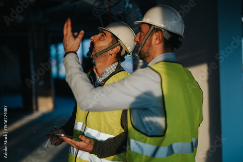 Fototapeta Two construction supervisors wearing safety vests and helmets inspecting a building site with intent