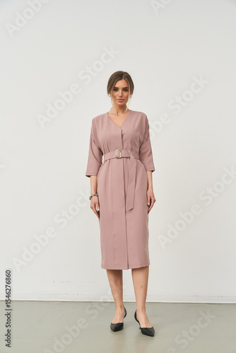 Elegant woman stands in minimalist style, wearing a belted knee-length dress with three-quarter sleeves and black shoes. Her hair is styled neatly, complementing her poised posture