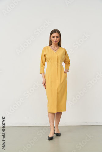 Woman in a long, yellow dress with three-quarter sleeves, featuring a v-neck and hand in pocket. She stands confidently in a minimalist setting, wearing black shoes, with straight hair