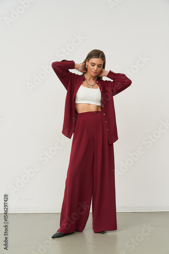 Young woman in a burgundy pantsuit with wide-leg trousers, wearing a white crop top underneath. She adjusts her hair with both hands, standing confidently on a beige floor