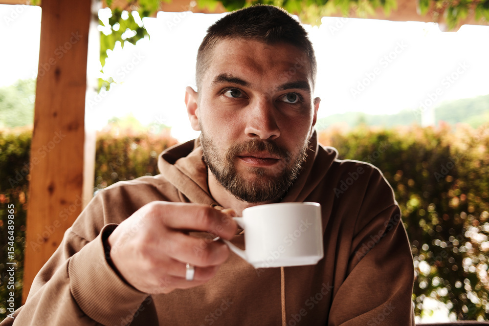 Bearded man in a brown hoodie drinking coffee at a traditional Serbian restaurant. Natural light and cozy wooden surroundings Bearded man in a brown hoodie drinking coffee at a traditional Serbian restaurant. Natural light and cozy wooden surroundings