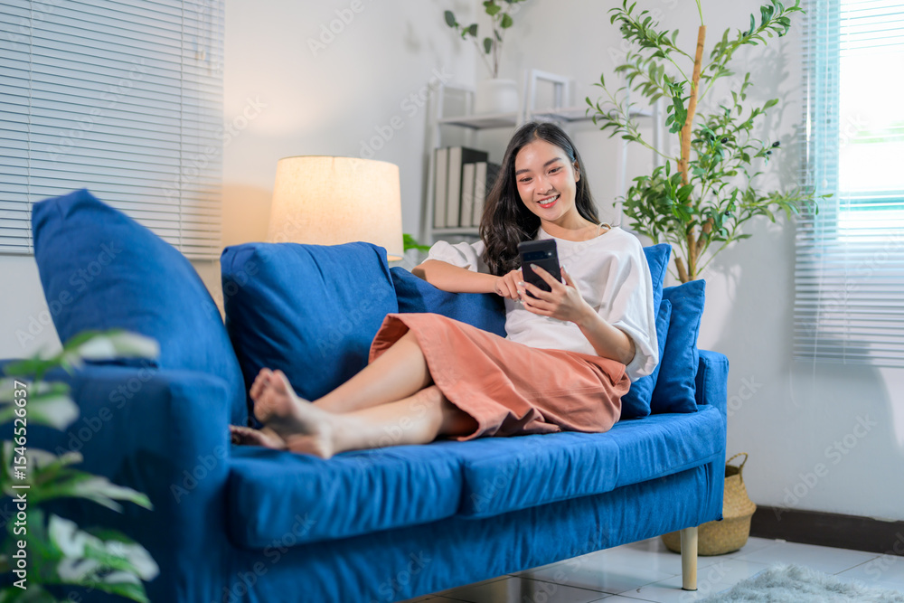 Obraz premium Happy asian woman using mobile phone while lying on comfortable sofa in living room at home