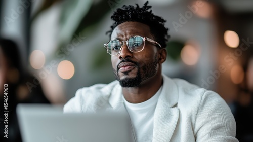 A contemplative man in a white blazer sits in a café, focused on his laptop, with a serene ambiance around him, reflecting a blend of work and casual lifestyle.
