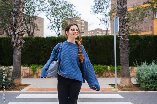 Happy student walking across the street on crosswalk while carrying a bag and smiling