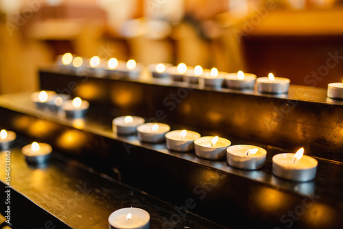 Fényképezés A row of tealight candles burns softly on a black metal stand, creating a peaceful and reflective ambiance often found in places of worship or memorials