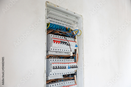 An open electrical panel embedded in a white wall, showing circuit breakers, blue, brown, and yellow green wires in an organized but unfinished state.