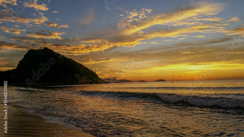 Praia do Leme, no Rio de Janeiro. Nascer do sol no mar, um dia com nuvens douradas, céu azul e ondas regulares. Morro do Forte à direita, montanhas ao fundo e algumas gaivotas.