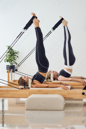 Side view of two sporty blonde women in black sportswear practicing pilates reformer, making shoulder stand exercise in modern studio.                                                           