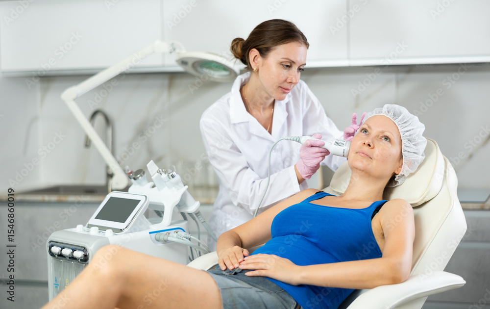 Obraz premium Young beautician with adult female client during hardware facial procedure in clinic. Concept of professional cosmetology and rejuvenation