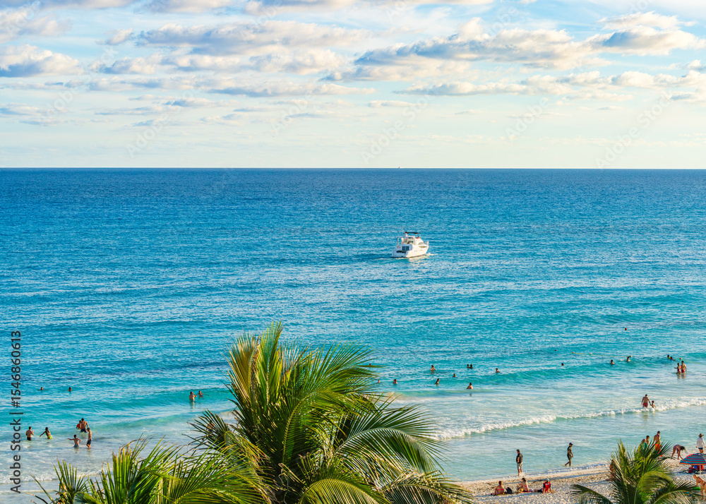Fototapeta premium People enjoying the beach and swimming in the clear blue waters of Cancun Hotel Zone with a boat cruising offshore under a partly cloudy sky High-quality photo.