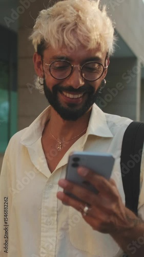 Vertical video, A young man with bleached blond hair and a beard smiles while walking down a city sidewalk and looking at his phone.