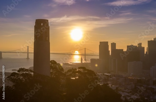 A scenic view of Coit Tower and the Bay Bridge at sunset in San Francisco, California, USA. The sun reflects off the water, creating a golden glow over the city.