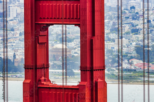 Photography Close-up of Golden Gate Bridge tower with Palace of Fine Arts in the background, San Francisco cityscape and boats on the bay visible through the iconic red structure