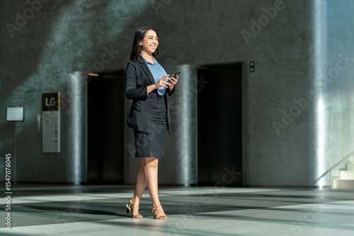 Smiling Asian businesswoman using smartphone in modern office lobby. Confident young professional walking with phone in elegant corporate interior
