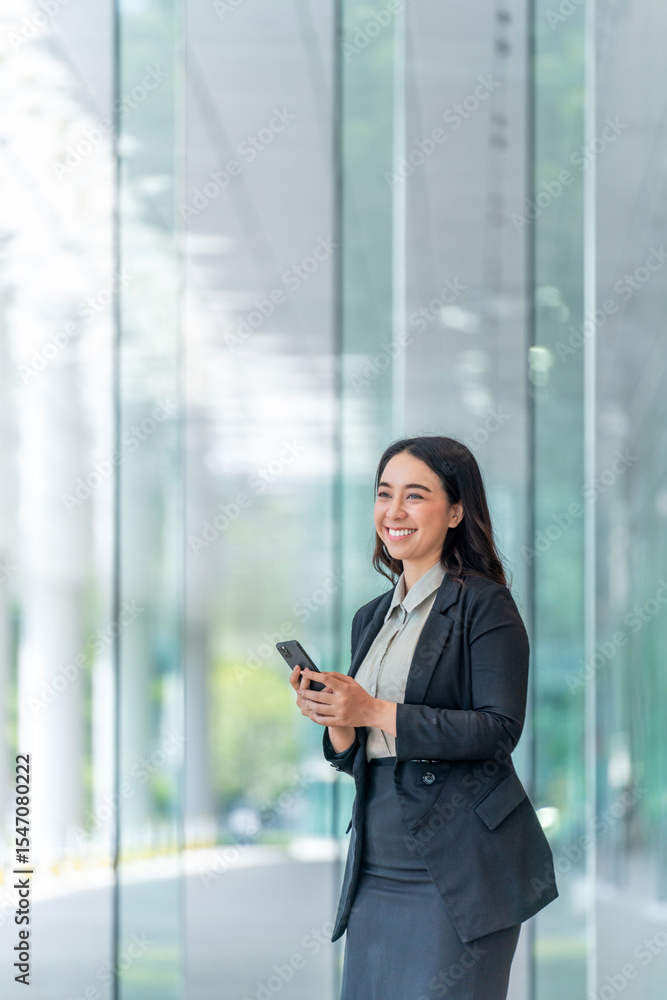 Fototapeta premium Smiling Asian businesswoman using smartphone in modern glass corridor. Confident woman in business attire standing indoors with mobile phone.