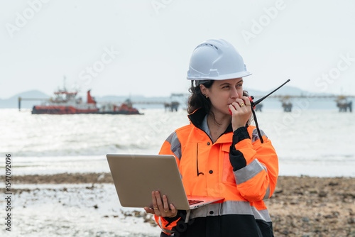 Female maritime engineer coordinating with a tugboat via radio at an offshore oil and gas terminal. Woman supervisor managing port operations with a laptop, with a ship in the background.