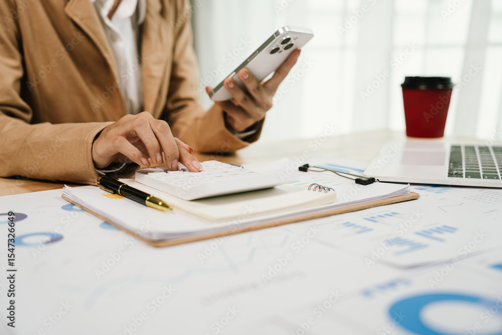 Fototapeta premium A dedicated professional analyzes charts on a clipboard with a pen, surrounded by technology like a laptop and smartphone, amidst a bustling work environment.