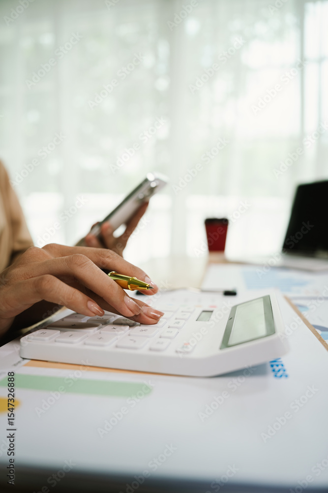 Fototapeta premium Close-up of businesswoman analyzing chart on clipboard with pen, surrounded by technology such as laptop and smartphone, in busy work environment.
