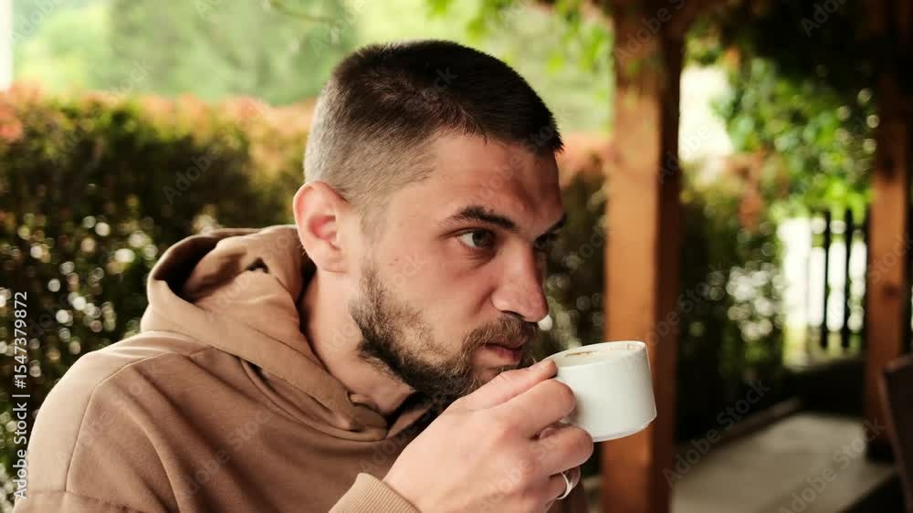 Bearded man in a brown hoodie drinking coffee at a traditional Serbian restaurant. Natural light and cozy wooden surroundings Bearded man in a brown hoodie drinking coffee at a traditional Serbian restaurant. Natural light and cozy wooden surroundings