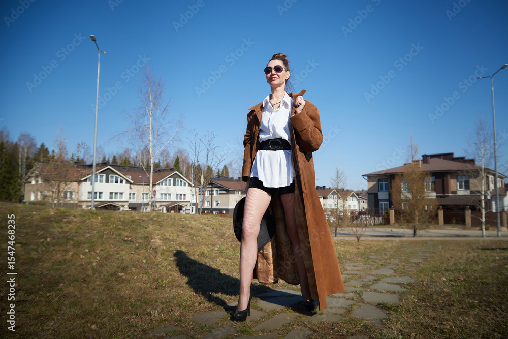 Fototapeta premium Confident woman in stylish outfit poses outdoors against modern homes in bright daylight