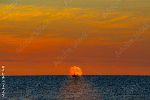 Small fishing vessel against the moon in the evening sea. Romantic landscape with a working boat at the beginning of the night.
