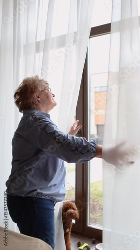 Senior woman standing by window at home, opening curtains to look outside and check weather, indoor, lifestyle