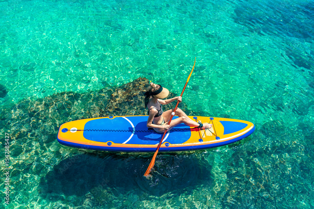 Fototapeta premium Aerial-style shot of a woman sitting and paddling over incredibly clear, emerald-green water, revealing rocks below the surface. A stunning visual of Croatia’s natural beauty
