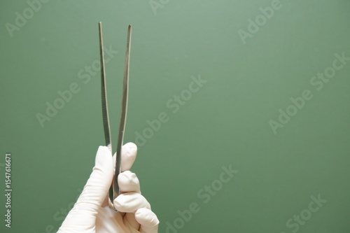 Gloved Hand Holding Metallic Tweezers Against Green Background