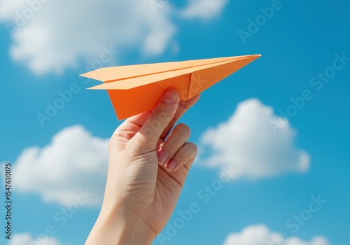 A hand holds an orange paper airplane against a clear blue sky with fluffy clouds, symbolizing dreams, freedom, and new ideas taking flight.