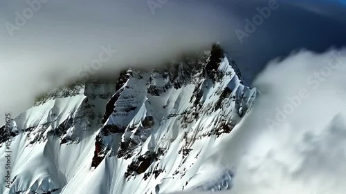 Snow and Wind Over Jagged Mountain Peak in Harsh Weather. Wide static shot of a rugged mountain peak shrouded in snow and low clouds. Wind whips snow across ridges. Raw, cinematic alpine scene