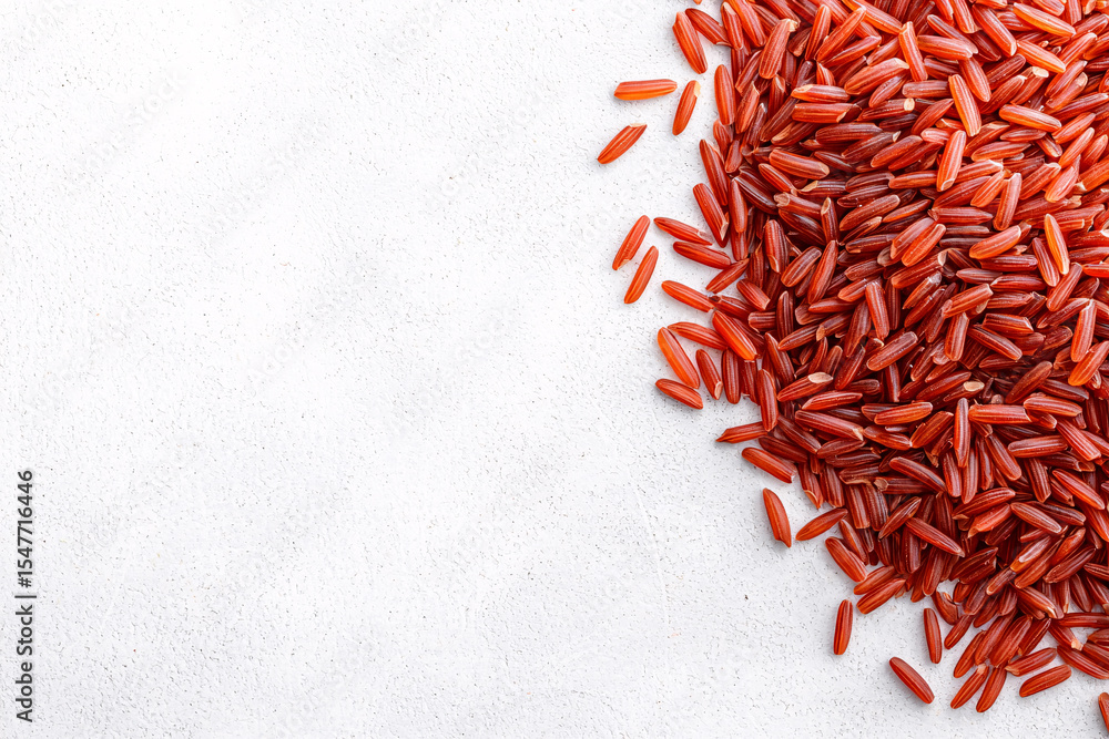 Fototapeta premium A scatte of uncooked red rice grains rests on a textured white surface with empty space left.