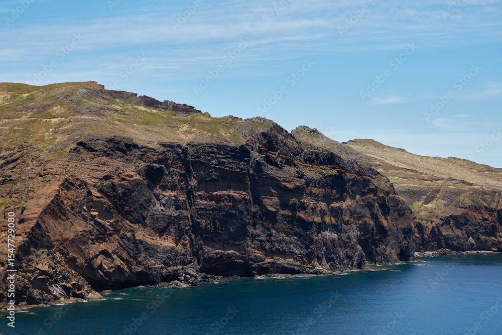 Fototapeta premium Dramatic rugged coastal cliffs towering over the deep blue ocean under a clear sky with hikers on a trail