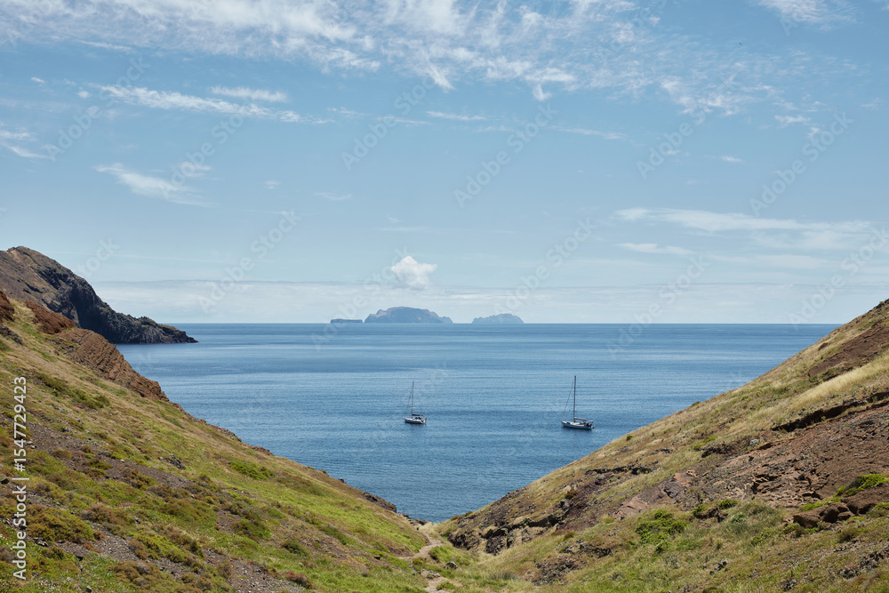 Fototapeta premium Scenic coastal landscape featuring sailboats on the sea, framed by natural rocky hills under a bright blue sky with distant islands on the horizon