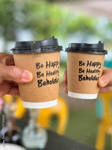 hands hold up takeaway coffee cups with black lids and motivational text that reads, “Be Happy, Be Healthy, Behold,” in an outdoor setting with blurred green trees and yellow chairs in the background.