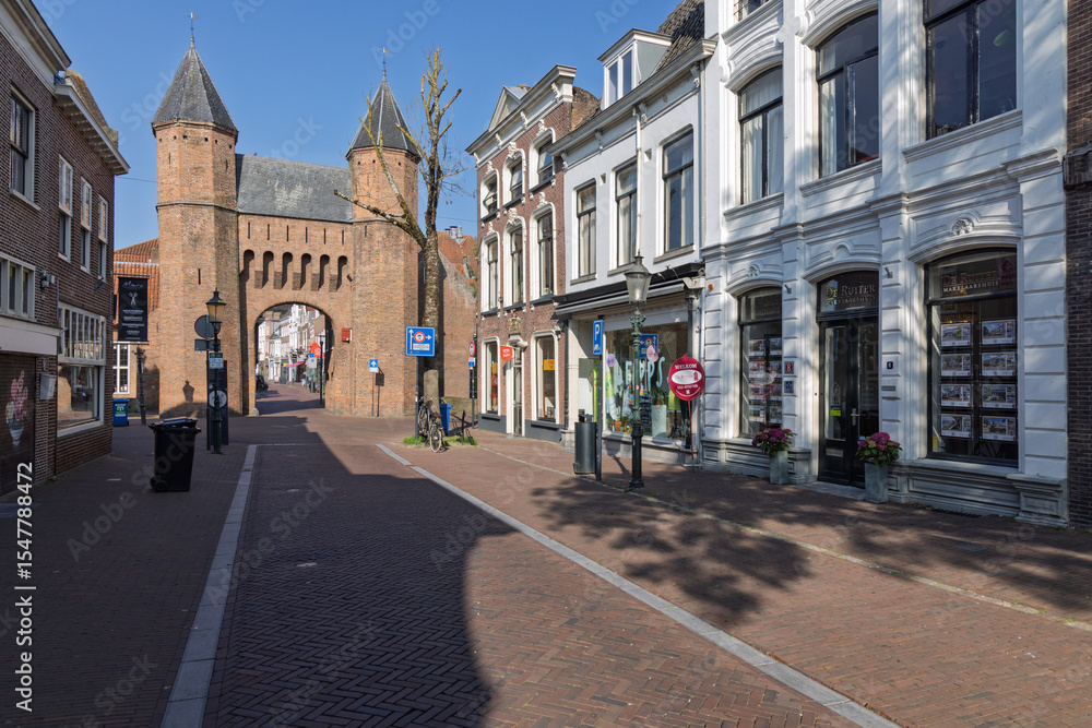 Naklejka premium The historic Kamperbinnenpoort gate in Amersfoort, Netherlands, frames a charming street with traditional Dutch architecture under a clear blue sky.