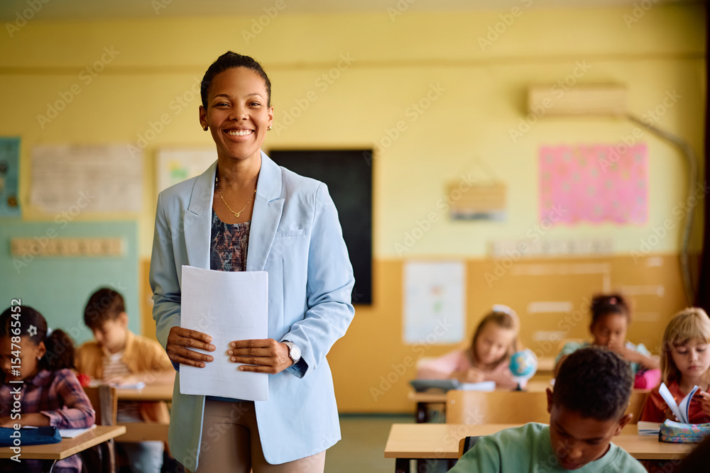 Fototapeta premium Happy black elementary school teacher among her students in classroom looking at camera.