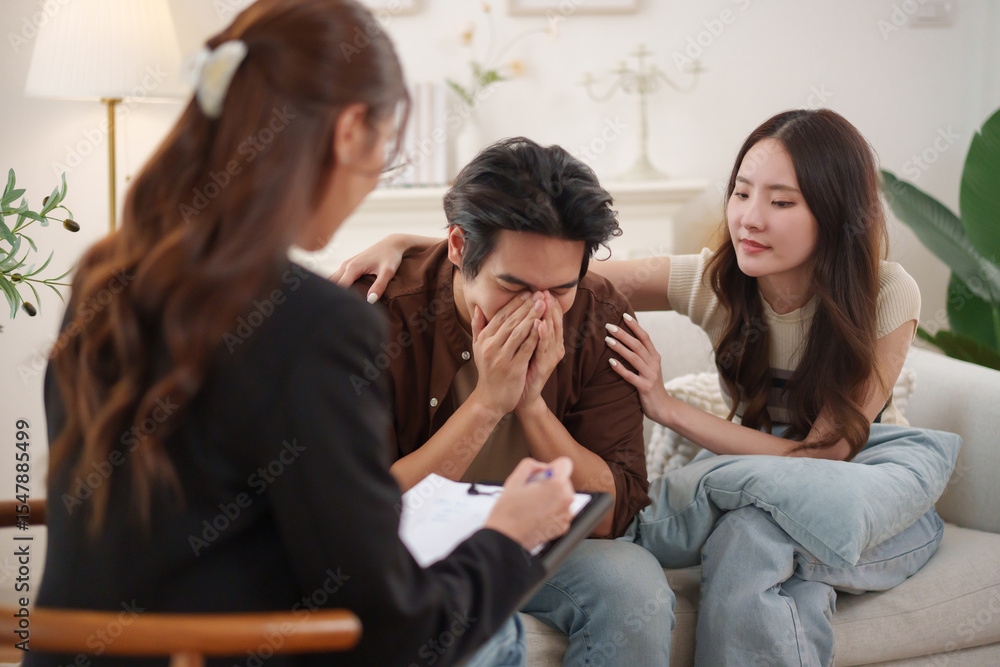 Young Asian man leaning forward in distress while being comforted by his wife during a counseling session with a female therapist. Young Asian man leaning forward in distress while being comforted by his wife during a counseling session with a female therapist.