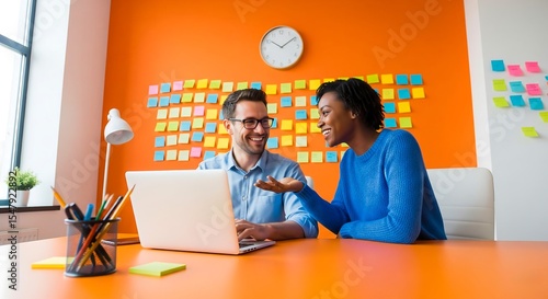 Two colleagues working together on laptop in bright office space with sticky notes and clock on the wall