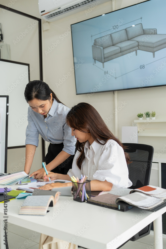Obraz premium Two Asian women working together in a bright, modern studio.while using a laptop and smartphone. The creative workspace is filled with colorful samples for seasonal palette selection.